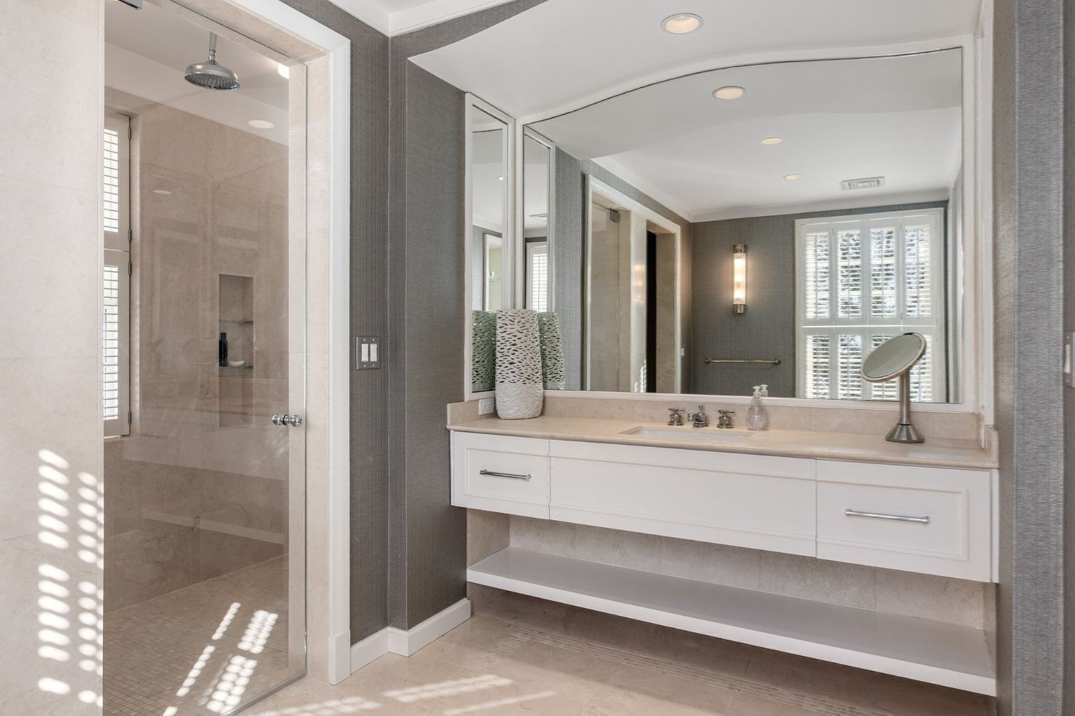 This primary bathroom features a sleek, floating white vanity with a light-toned stone countertop and a large, arched mirror. To the left, a glass-enclosed walk-in shower is visible, complete with a rainfall showerhead and built-in shelving. The room is finished with textured dark gray wallpaper and light-colored tile flooring, creating a sophisticated and modern aesthetic.