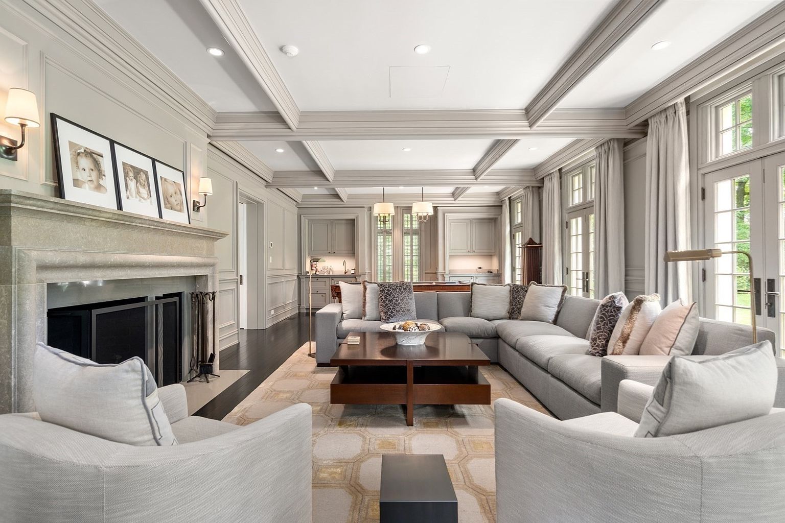 This elegant living room features a coffered ceiling and a neutral color palette, centered around a large stone fireplace with a prominent mantel. A spacious sectional sofa and two armchairs are arranged on a patterned area rug, creating a comfortable seating area, while a wet bar is visible in the background. The room is bathed in natural light from tall windows and glass doors, offering a sophisticated and inviting atmosphere.