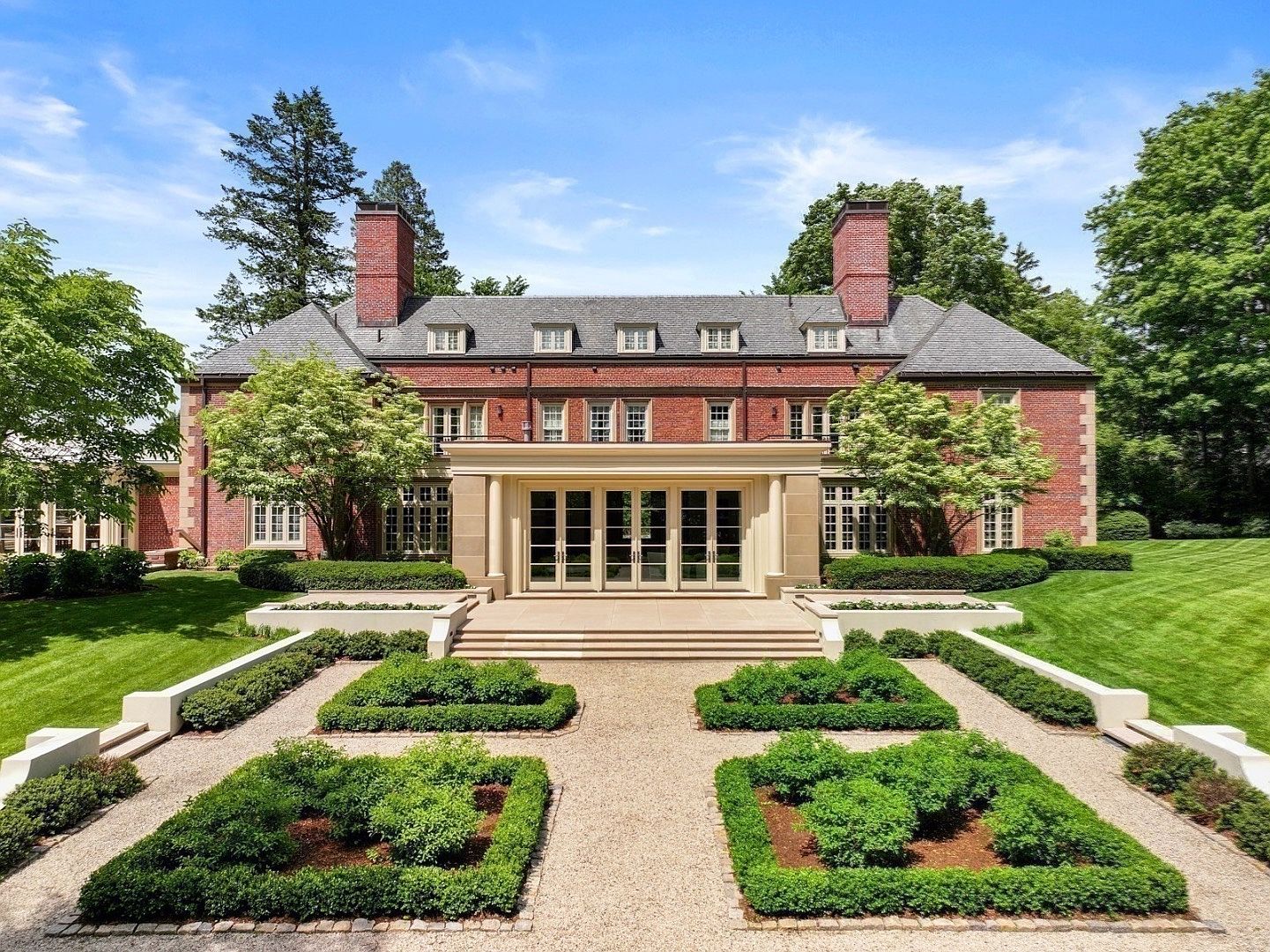 This grand brick estate features a symmetrical rear facade with a prominent central portico leading to a stone terrace. The meticulously manicured formal garden in the foreground includes geometric boxwood hedges and gravel pathways, creating a classic, stately aesthetic. The perspective is a wide, eye-level shot that captures the impressive scale of the architecture against a clear blue sky.