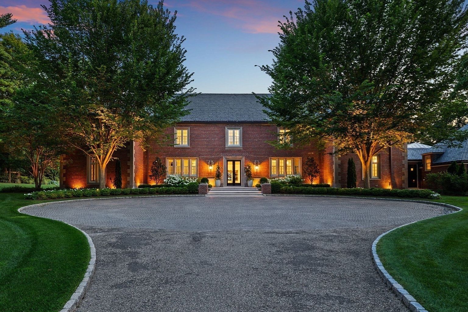 This stately brick residence is captured from a front-facing perspective, showcasing a symmetrical facade framed by two large, mature trees. The home features a classic architectural style with multi-pane windows, a central entryway with a dark door, and warm exterior lighting that highlights the masonry and landscaping. A circular gravel driveway leads directly to the front steps, creating a grand and welcoming approach to the property.