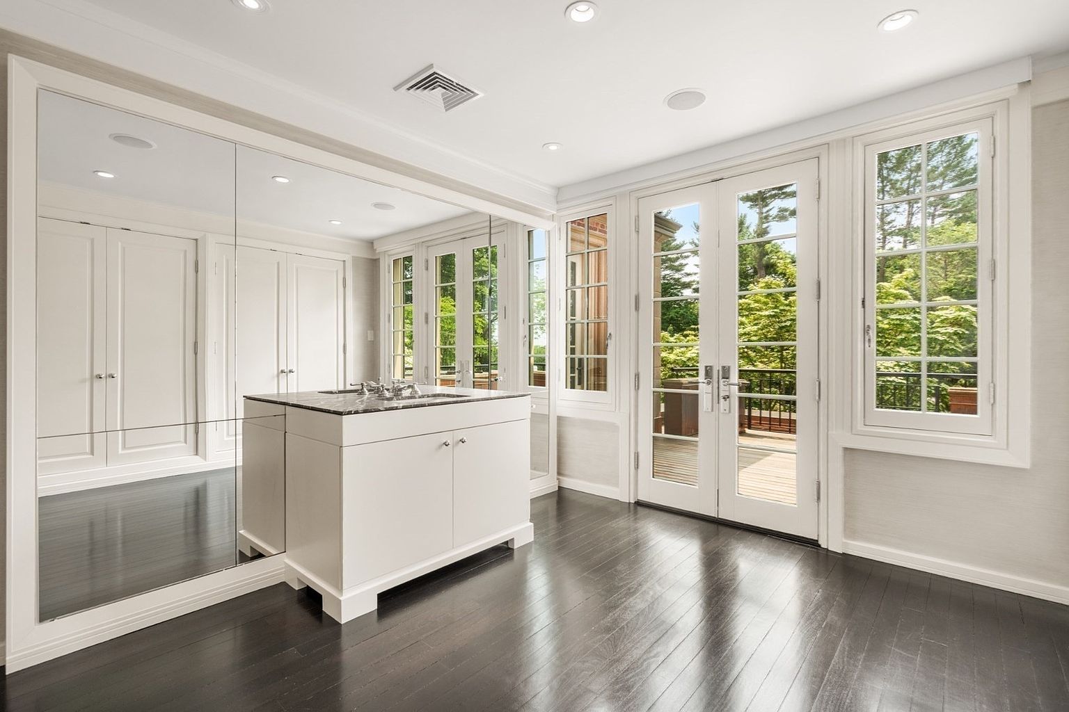 This sophisticated walk-in closet features dark hardwood flooring and a central island topped with a dark stone surface. A large mirrored wall reflects the space, enhancing the sense of depth, while white cabinetry provides ample storage. The room is brightened by natural light streaming through a set of French doors and adjacent windows that offer a view of the lush greenery outside.