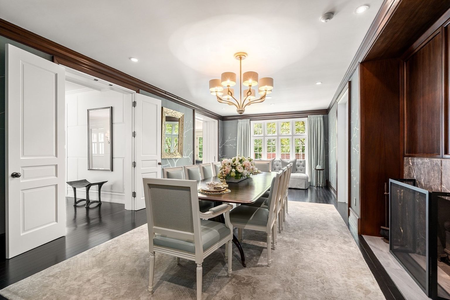 This elegant dining room features a long wooden table surrounded by upholstered chairs, set beneath a sophisticated multi-arm chandelier. The space is defined by light blue wallpaper with delicate white branch patterns, dark wood flooring, and a stone-clad fireplace surround on the right. A large window at the far end of the room provides natural light, complemented by a neutral area rug that anchors the formal seating arrangement.