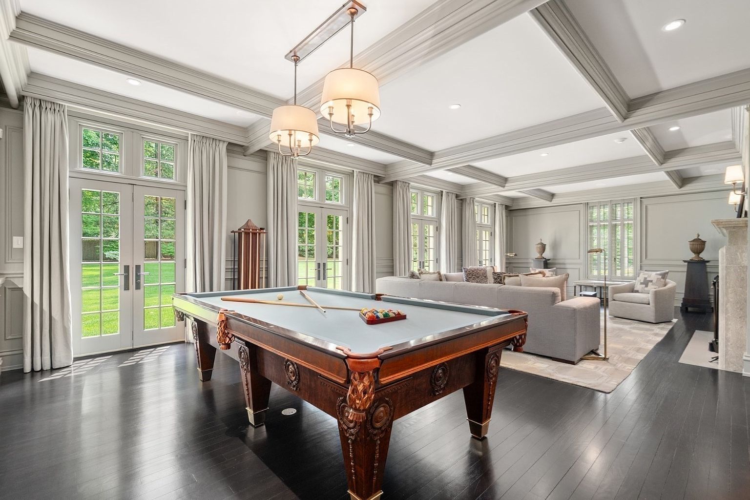 This spacious, open-concept room features a classic billiards table as a central focal point, set against a backdrop of dark hardwood flooring and light-toned wall paneling. The space transitions into a comfortable seating area with a large sofa and armchairs, illuminated by elegant pendant lighting and natural light streaming through multiple sets of French doors. The coffered ceiling and traditional architectural details contribute to a sophisticated and inviting atmosphere.