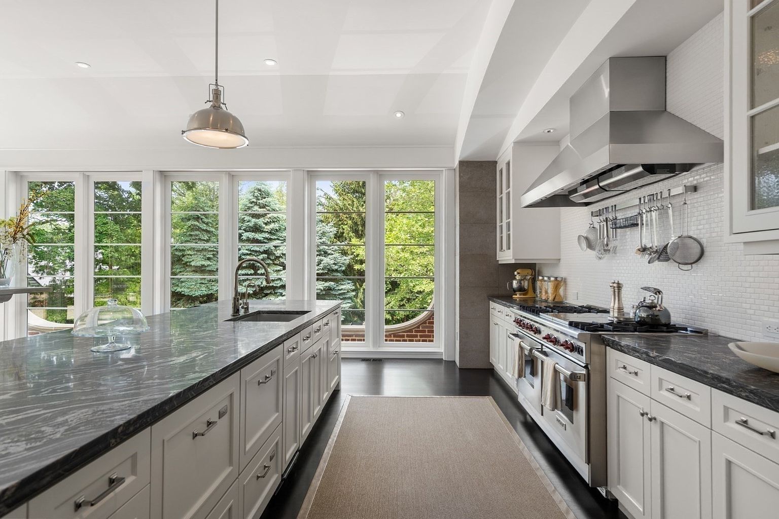 This bright, high-end kitchen features a large central island with a dark stone countertop and white cabinetry, complemented by a matching stone-topped perimeter workspace. A professional-grade stainless steel range with a matching hood serves as the focal point, while a row of tall windows provides abundant natural light and views of the surrounding greenery. The space is finished with dark flooring and a neutral runner, creating a sophisticated and airy atmosphere.