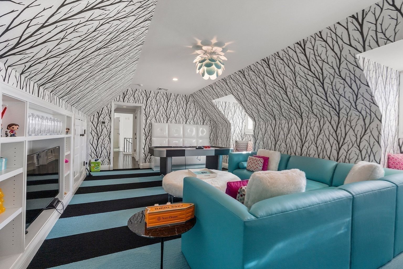 This vibrant attic game room features a striking tree-branch patterned wallpaper that extends across the sloped ceilings and walls. The space is furnished with a bright turquoise sectional sofa, a white circular ottoman, and a game table, all set against a bold black and blue striped carpet. A unique, multi-petaled light fixture hangs from the center of the room, adding a whimsical touch to this playful entertainment area.
