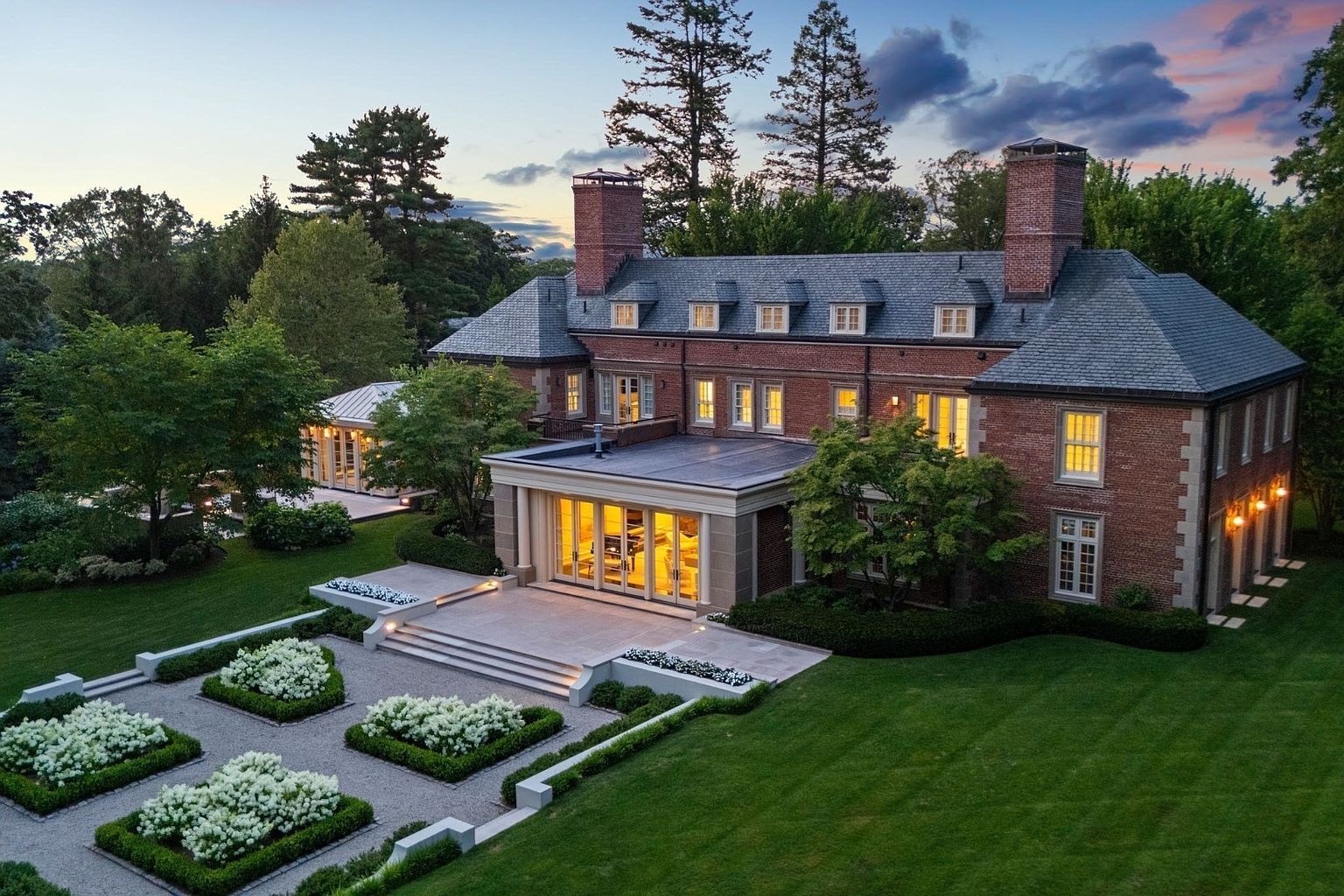 This aerial perspective captures a stately brick manor house at dusk, showcasing its classic architectural design and symmetrical facade. The property features a manicured formal garden with geometric boxwood hedges and white flowering shrubs, leading to a stone patio with a glass-enclosed sunroom. The warm glow from the interior lights contrasts beautifully with the twilight sky, highlighting the home's elegant proportions and expansive, well-maintained lawn.