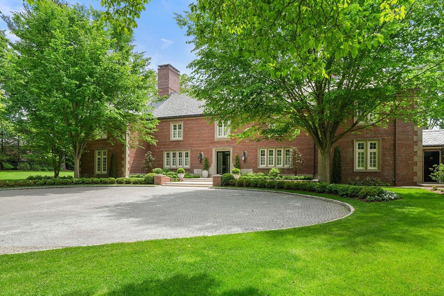 This stately brick residence features a symmetrical facade with a prominent central entryway and a circular cobblestone driveway. The home is surrounded by mature trees and manicured landscaping, creating a private and elegant curb appeal. The classic architectural style is complemented by multi-pane windows and a slate-style roof, presenting a refined and timeless aesthetic.