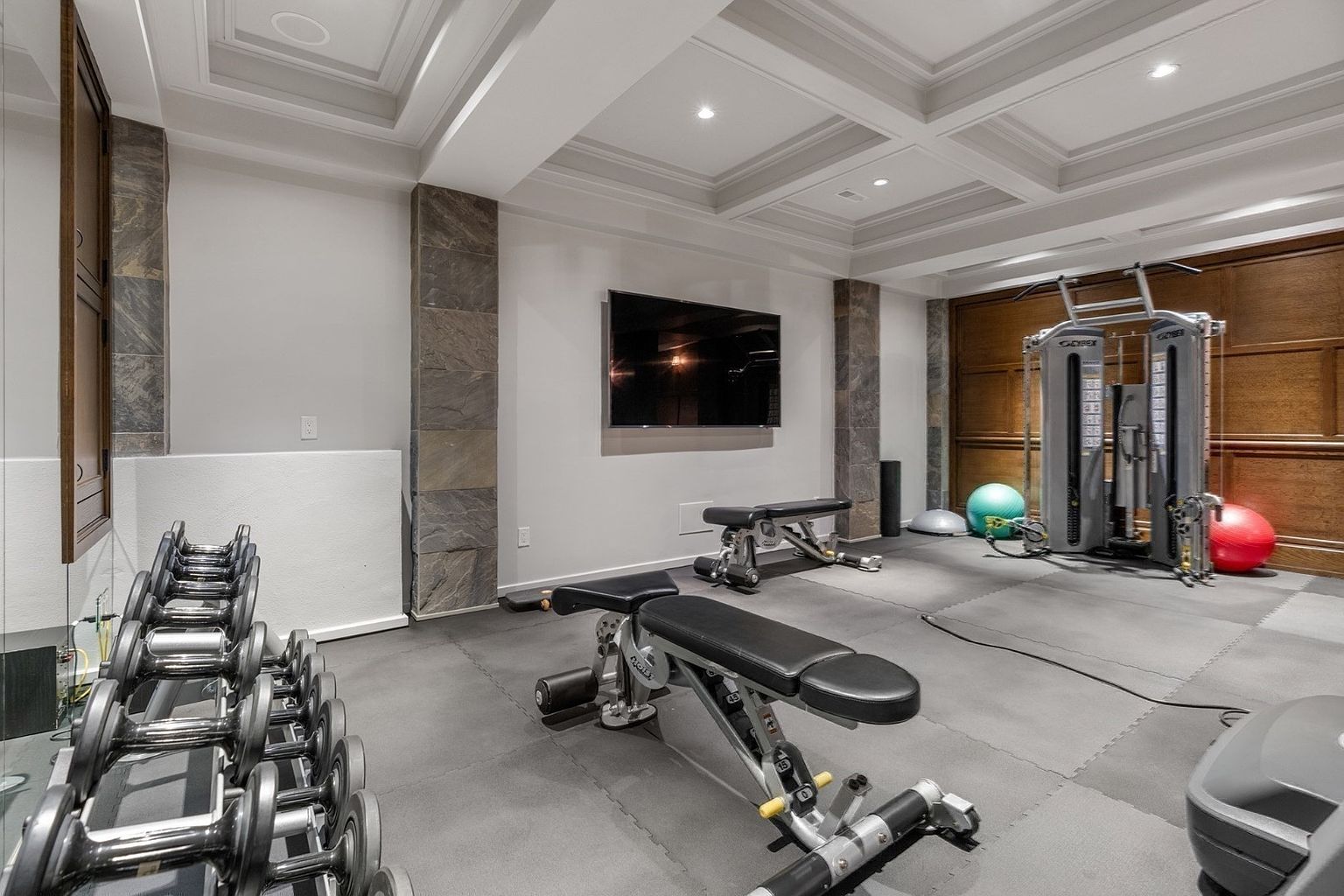 This well-equipped home gym features a professional-grade cable machine, multiple adjustable workout benches, and a rack of dumbbells. The space is finished with gray interlocking floor mats, stone-clad support columns, and a coffered ceiling with recessed lighting. A large flat-screen television is mounted on the central wall, creating a functional and modern fitness environment.
