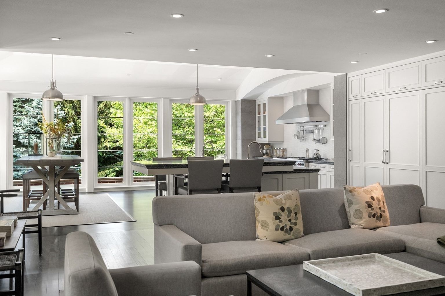 This open-concept living space seamlessly integrates a comfortable seating area with a kitchen and dining zone. The room features a neutral color palette, dark wood flooring, and large windows that provide abundant natural light and views of the surrounding greenery. The kitchen is equipped with stainless steel appliances and a central island, creating a functional and inviting atmosphere for entertaining.