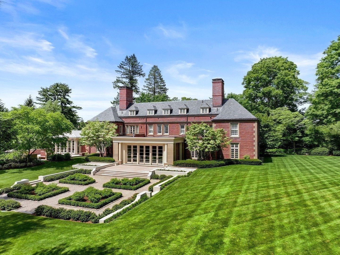 This grand brick estate is captured from an elevated perspective, showcasing its symmetrical Georgian-inspired architecture and expansive, meticulously manicured grounds. A formal parterre garden with geometric hedges sits in the foreground, leading to a stone terrace and a central sunroom addition with large glass doors. The property is surrounded by lush, mature trees under a bright blue sky, creating a serene and stately atmosphere.