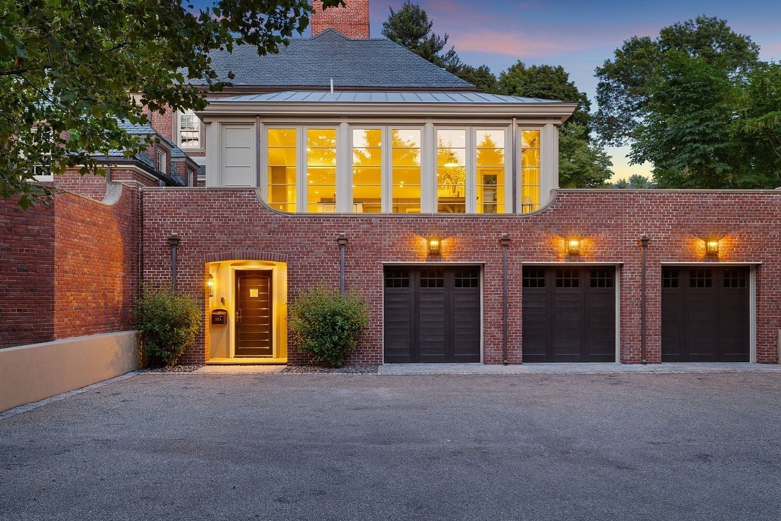This elegant brick exterior features a three-car garage with dark, paneled doors and warm wall-mounted lighting. Above the garage, a sun-drenched room with large windows creates a striking architectural contrast against the traditional masonry. The scene is captured at dusk, highlighting the inviting glow from the entryway and the upper-level windows against the twilight sky.