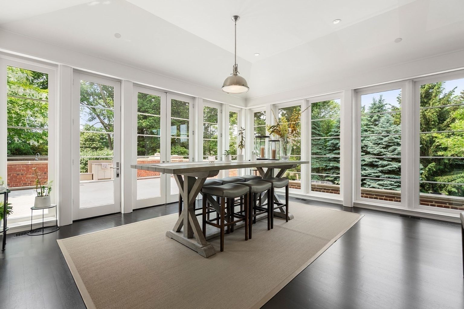 This bright sunroom features a long, rustic-style wooden dining table centered on a neutral area rug, surrounded by dark-toned bar stools. The space is defined by expansive, floor-to-ceiling windows and glass doors that provide abundant natural light and panoramic views of the surrounding greenery. A single industrial-style pendant light hangs above the table, creating a clean, airy atmosphere that seamlessly connects the interior with the outdoors.