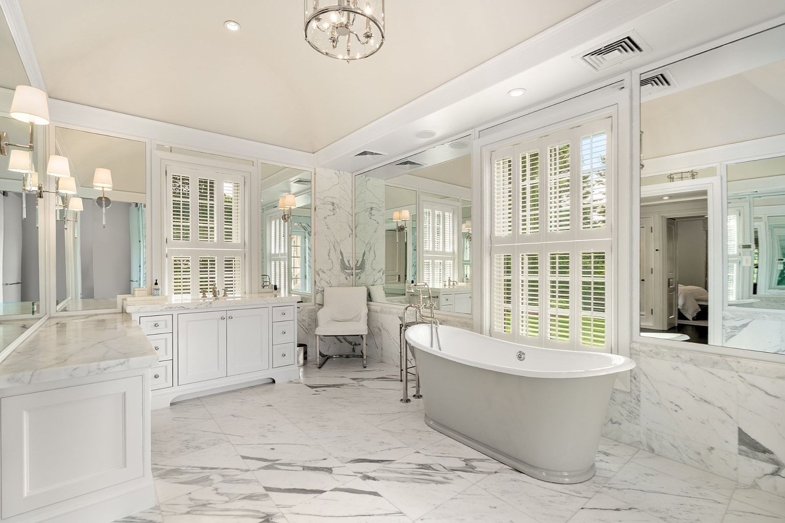 This luxurious primary bathroom features a freestanding soaking tub as its centerpiece, set against a backdrop of white stone walls and large mirrors. The space is illuminated by a decorative chandelier and wall-mounted sconces, creating a bright and elegant atmosphere. White cabinetry with stone countertops and patterned stone flooring complete the sophisticated, high-end aesthetic.