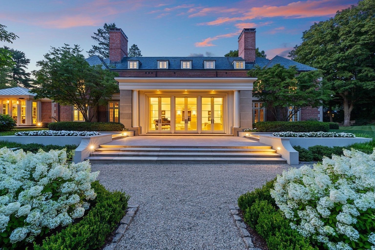 This elegant brick estate is captured from a rear perspective at twilight, showcasing a symmetrical architectural design with a prominent central portico. The home features a series of tall glass doors that open onto a stone terrace, which is framed by manicured hedges and lush white hydrangea bushes. The scene is illuminated by warm, inviting exterior lighting that highlights the stone steps and the structured landscaping, creating a sophisticated and serene atmosphere.