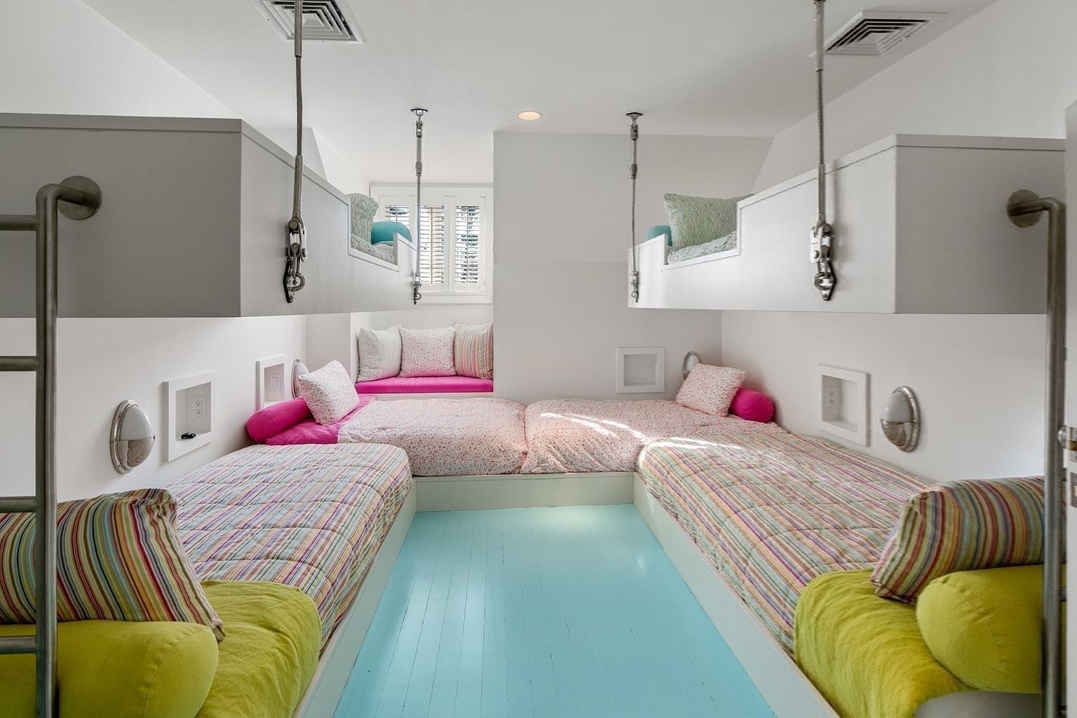 This charming, symmetrical bedroom features four built-in beds arranged in a U-shape, with two lower beds and two suspended upper bunks. The room is accented by a vibrant light blue painted floor, striped bedding, and a cozy window seat at the back. The space is bright and airy, utilizing white walls and industrial-style metal hardware to create a playful yet organized sleeping area.