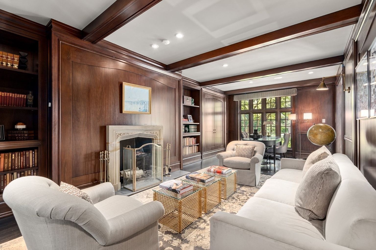 This sophisticated study features rich wood-paneled walls, built-in bookshelves, and a classic stone fireplace surround that serves as a focal point. The room is furnished with a plush sofa, two armchairs, and a pair of gold-toned coffee tables, all arranged on a patterned area rug. A cinematic perspective captures the depth of the room, leading the eye toward a set of large windows at the far end that provide natural light and a view of the outdoors.