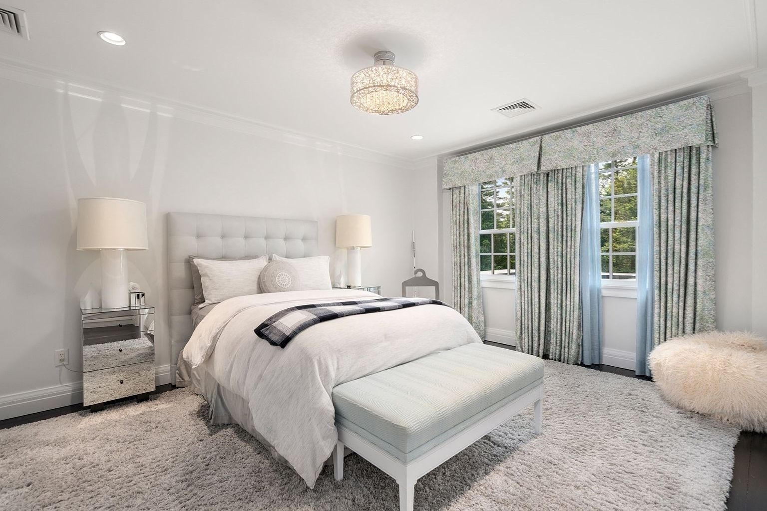 This bright and airy primary bedroom features a plush, tufted headboard and a neutral color palette accented by soft blue patterned curtains. A mirrored nightstand adds a touch of modern elegance, while a textured rug and a fluffy ottoman create a cozy, inviting atmosphere. The room is illuminated by a decorative chandelier and recessed lighting, offering a serene and polished aesthetic.