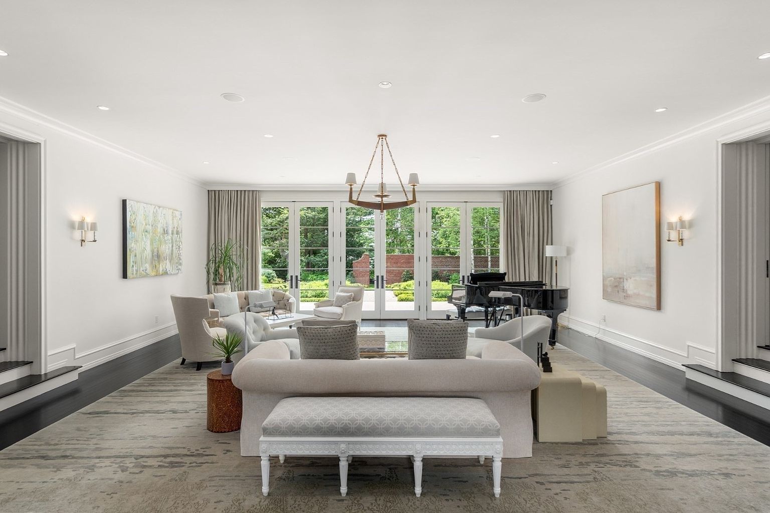 This elegant and spacious living room features a symmetrical layout centered around a plush sofa and a patterned bench, set upon a large, neutral-toned area rug. The room is anchored by dark hardwood floors and bright white walls, with a grand piano positioned near the rear wall. Large glass doors provide a clear view of the lush green garden outside, while a sophisticated chandelier hangs centrally to illuminate the refined, contemporary space.