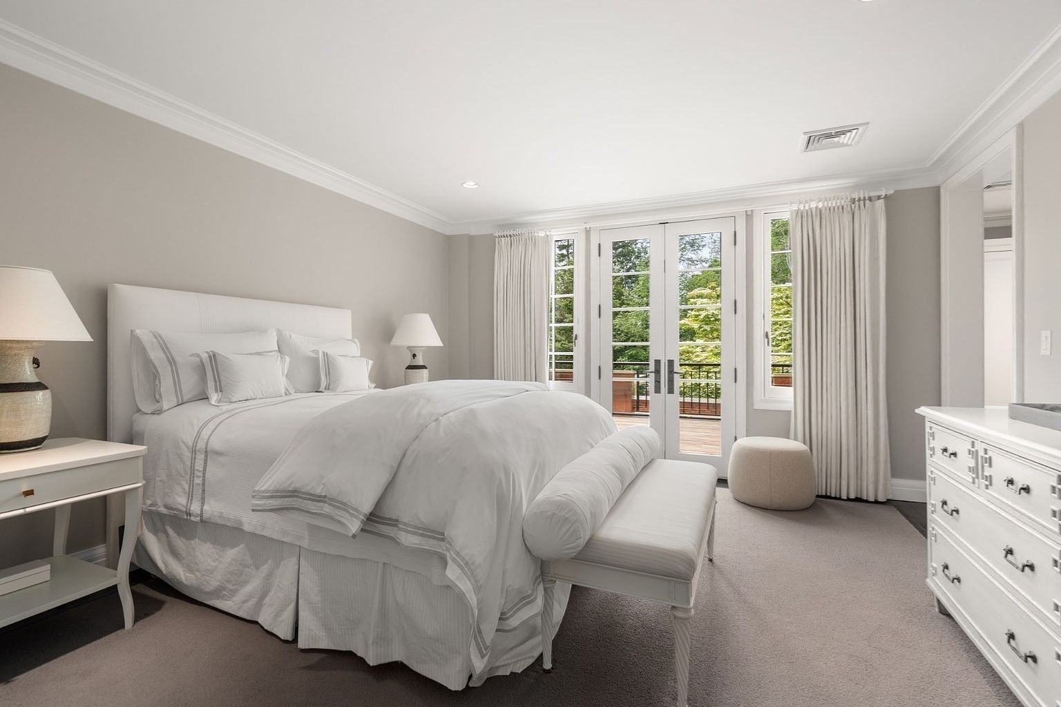 This serene primary bedroom features a neutral color palette with soft gray walls and plush carpeting, creating a tranquil atmosphere. A large, upholstered bed serves as the focal point, complemented by matching nightstands and a white dresser. Tall glass doors open to a private outdoor deck, allowing ample natural light to fill the room.