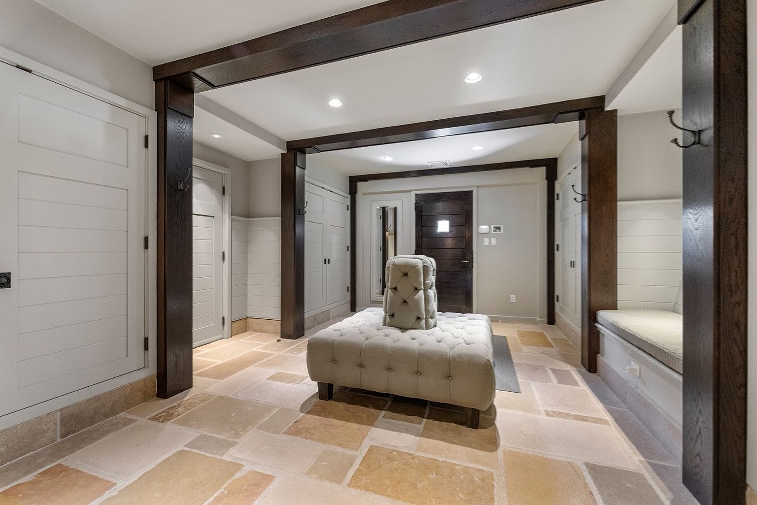 This spacious entryway features a neutral stone tile floor and dark wood structural beams that frame the transition between areas. A central tufted ottoman with a vertical backrest serves as a focal point, while built-in cabinetry and a bench with hooks provide functional storage. The space is illuminated by recessed lighting, creating a warm and inviting atmosphere for guests.