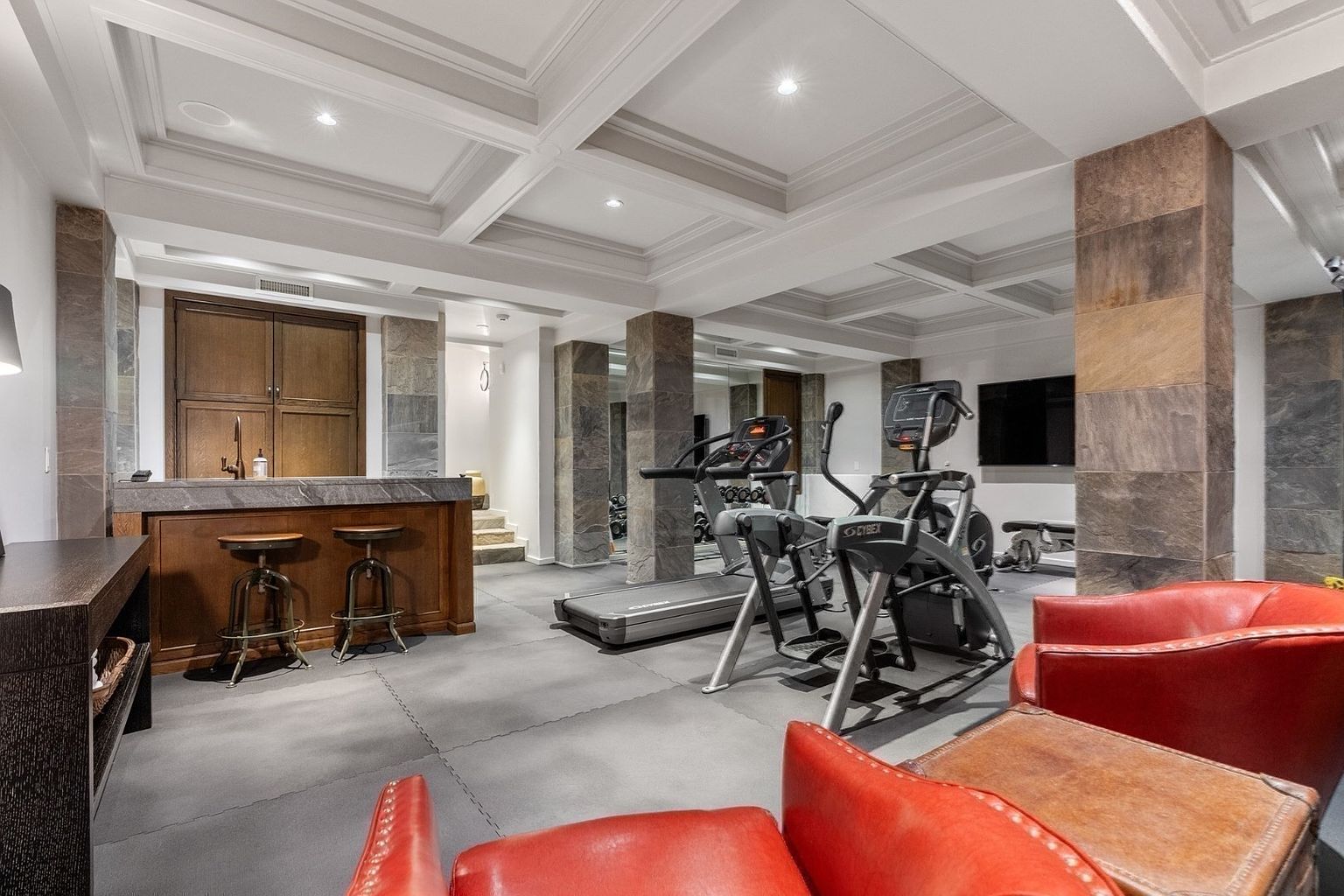This spacious home gym features a dedicated workout area equipped with a treadmill and an elliptical machine, set against a backdrop of stone-clad support columns. To the side, a built-in bar area with stools provides a relaxed space for post-workout recovery, while red leather armchairs offer comfortable seating in the foreground. The room is finished with a coffered ceiling and dark, textured flooring, creating a sophisticated and functional multi-purpose environment.