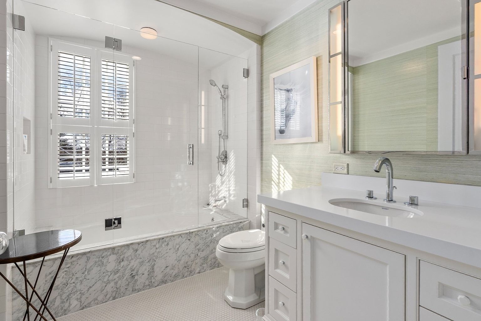 This bright and elegant bathroom features a combination tub and shower enclosure with a glass partition and white subway tile walls. A vanity with a white countertop and a single sink sits against a wall covered in textured, light-toned wallpaper, complemented by a large medicine cabinet mirror. The space is finished with a stone-fronted tub apron and small-scale floor tiles, creating a clean and sophisticated aesthetic.