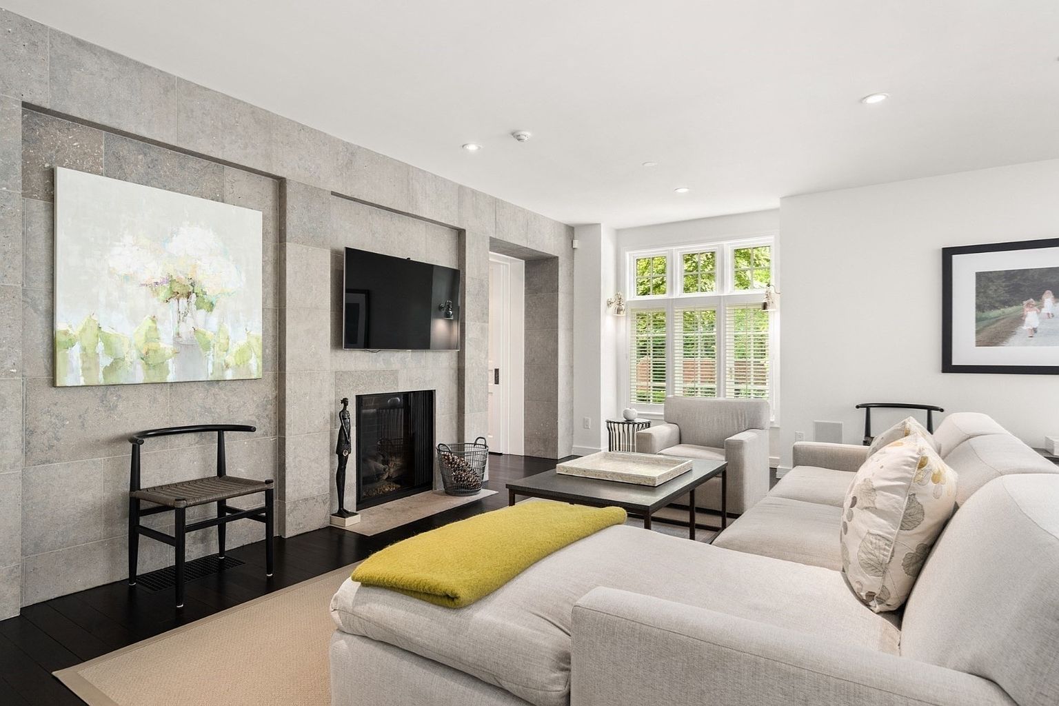 This bright and inviting living room features a prominent stone-clad chimney breast that serves as a focal point, housing a television and a fireplace. A large, comfortable sectional sofa with a yellow throw blanket anchors the seating area, complemented by a central coffee table and a minimalist chair. The space is finished with dark wood flooring and large windows with plantation shutters that allow natural light to fill the room.