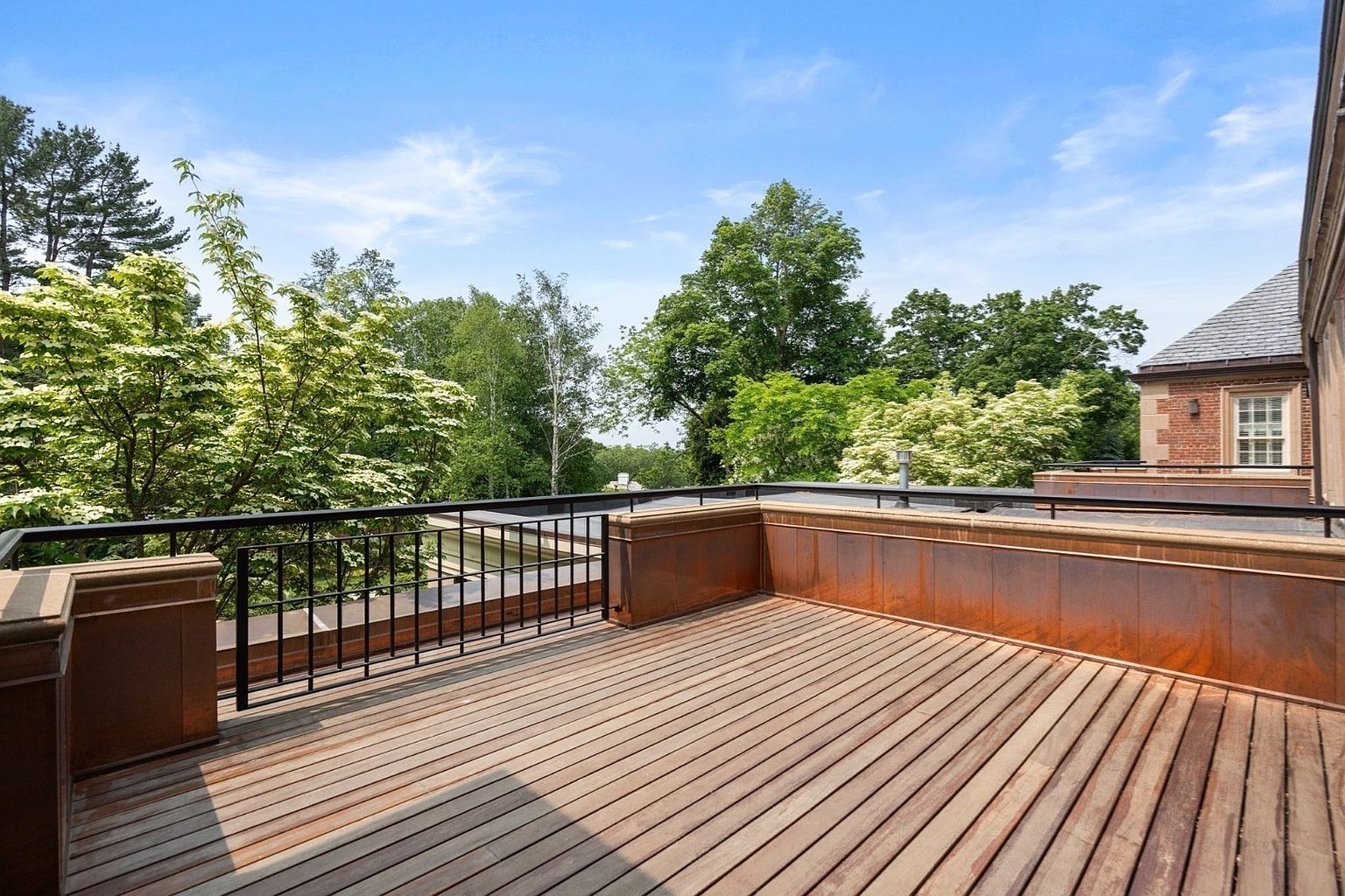 This elevated wooden deck features a clean, slatted floor design enclosed by a combination of metal railings and copper-toned panels. The space offers an expansive, open-air perspective overlooking a lush canopy of mature trees under a clear blue sky. It provides a serene and private outdoor retreat, seamlessly blending architectural structure with the surrounding natural landscape.