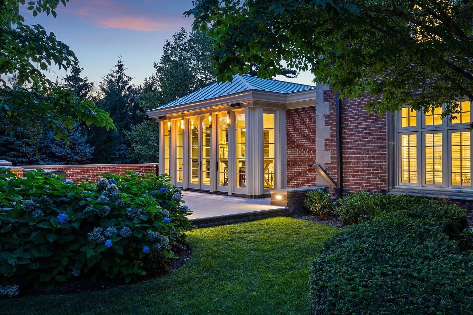 This evening view captures a sophisticated brick residence featuring a sunroom addition with large, multi-pane windows and a metal roof. A manicured lawn and lush hydrangea bushes frame the stone patio, creating a serene and inviting outdoor living space. The warm glow from the interior lights contrasts beautifully with the twilight sky, highlighting the home's elegant architectural details.
