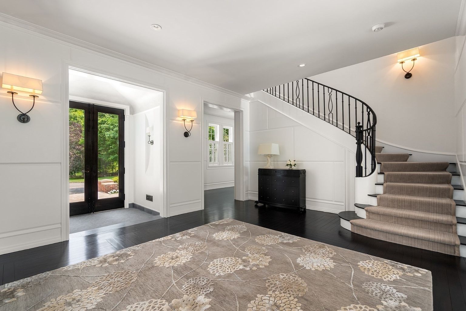 This elegant foyer features dark hardwood flooring contrasted by crisp white walls with classic wainscoting. A grand curved staircase with a black iron railing and a neutral carpet runner serves as the focal point, while a large, patterned area rug anchors the space. The entryway is illuminated by warm wall sconces and a view through the double glass doors that leads to the exterior.