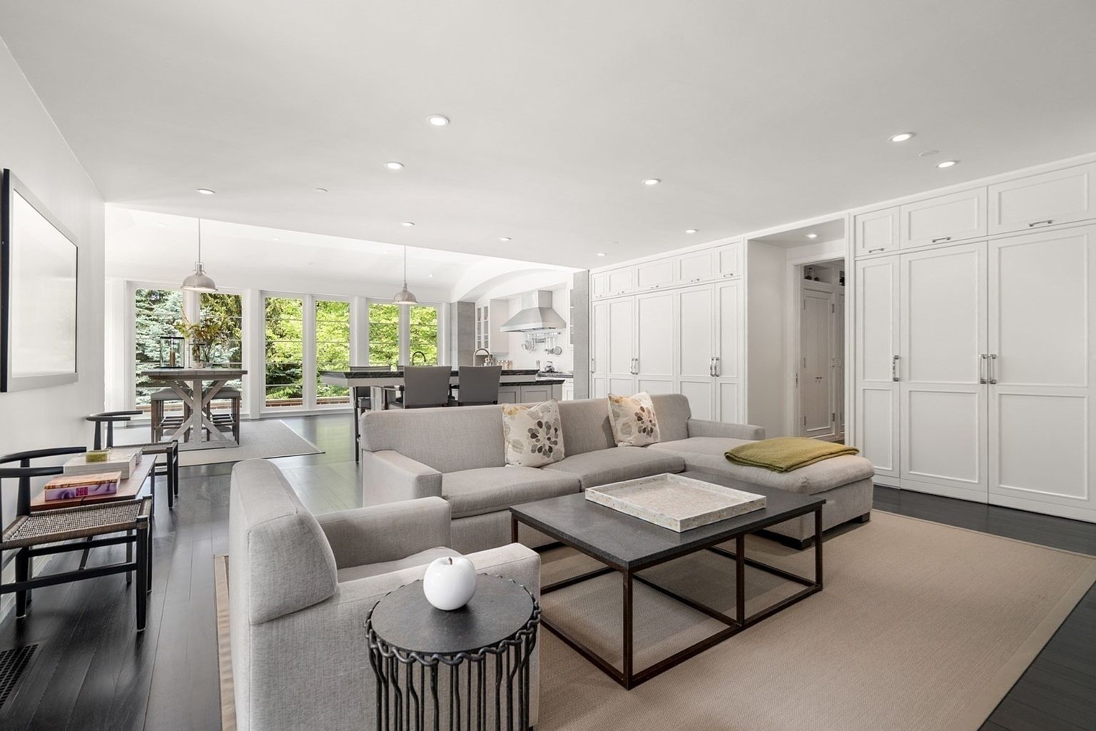 This open-concept living space features a neutral color palette with a large sectional sofa and a matching armchair arranged around a dark metal-framed coffee table. The room flows seamlessly into a dining area and kitchen, illuminated by recessed lighting and large windows that provide a view of the lush greenery outside. The dark wood flooring contrasts with the crisp white walls and extensive built-in cabinetry, creating a sophisticated and airy atmosphere.