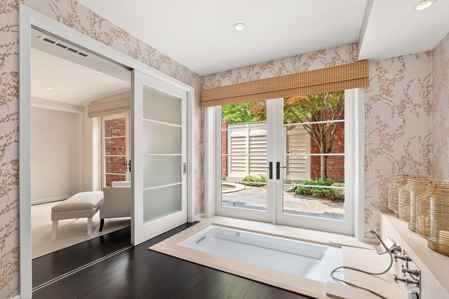 This luxurious primary bathroom features a deep soaking tub set into a light-toned stone deck, positioned directly in front of large glass doors that open to a private garden view. The walls are adorned with elegant floral-patterned wallpaper, complemented by dark wood flooring and a sliding frosted-glass door leading to an adjacent room. The space exudes a serene, spa-like atmosphere with natural light streaming in through the garden-facing windows.
