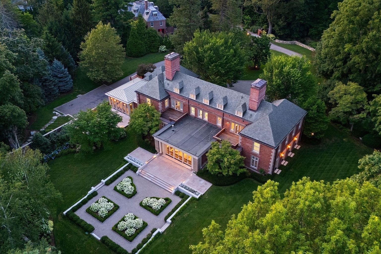 This high-angle aerial view captures a grand, multi-story brick estate nestled within a lush, mature landscape. The property features a formal garden with geometric hedges and white flowering shrubs, a stone patio, and a sunroom extension, all set against a backdrop of dense, verdant trees. The classic architectural style is highlighted by the symmetrical brick facade, slate-style roof, and prominent chimneys, creating an impression of timeless elegance and privacy.