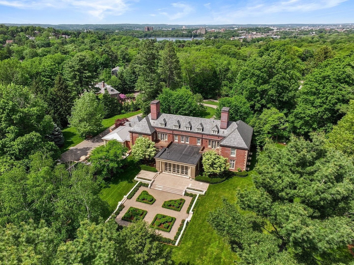 This high-angle aerial view captures a grand, multi-story brick estate nestled within a lush, mature forest. The property features a formal geometric garden with manicured hedges and stone pathways leading to a flat-roofed sunroom extension. The expansive grounds are surrounded by dense greenery, providing a sense of privacy and seclusion in a serene, park-like setting.