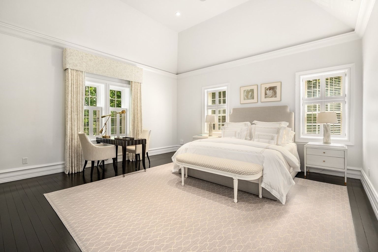 This spacious primary bedroom features high vaulted ceilings and dark hardwood floors, creating an airy yet grounded atmosphere. The room is anchored by a neutral-toned upholstered bed, flanked by matching white nightstands and lamps, with a patterned bench at the foot of the bed. To the left, a dedicated workspace with a sleek desk and two chairs is positioned near large windows dressed in elegant floor-length drapery.
