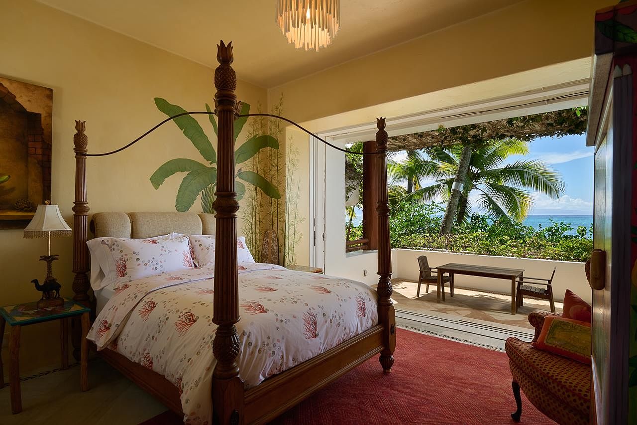 This primary bedroom features a four-poster bed with decorative pineapple finials, complemented by tropical-themed wall art and a view of the ocean. The room opens onto a balcony with outdoor seating, creating a seamless indoor-outdoor living experience. The overall aesthetic is luxurious and relaxing, perfect for a vacation rental or high-end property listing.
