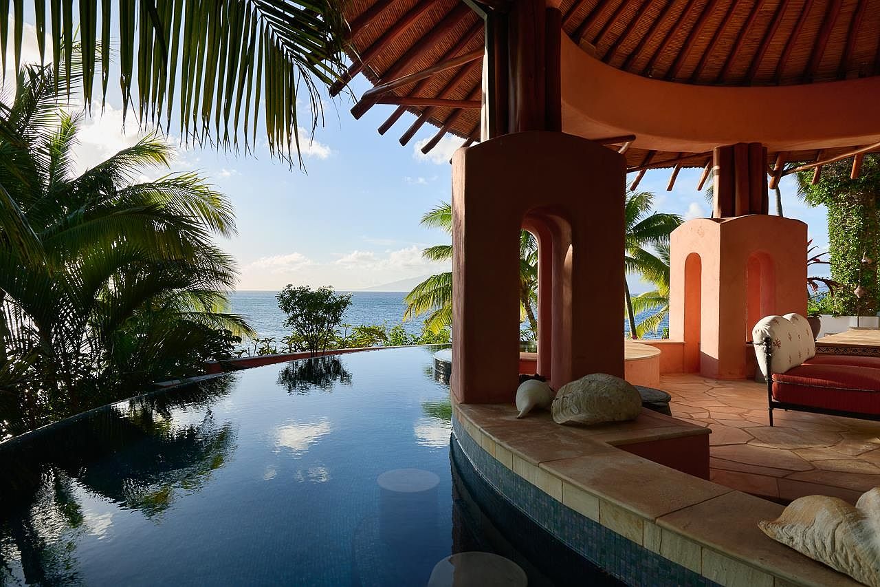 This image showcases a luxurious infinity pool overlooking the ocean, framed by lush tropical foliage and a unique architectural structure with arched openings. The pool's dark blue water contrasts beautifully with the light blue ocean and sky, creating a serene and inviting atmosphere. The architectural details and the seamless blend of indoor and outdoor spaces suggest a high-end resort or private villa.