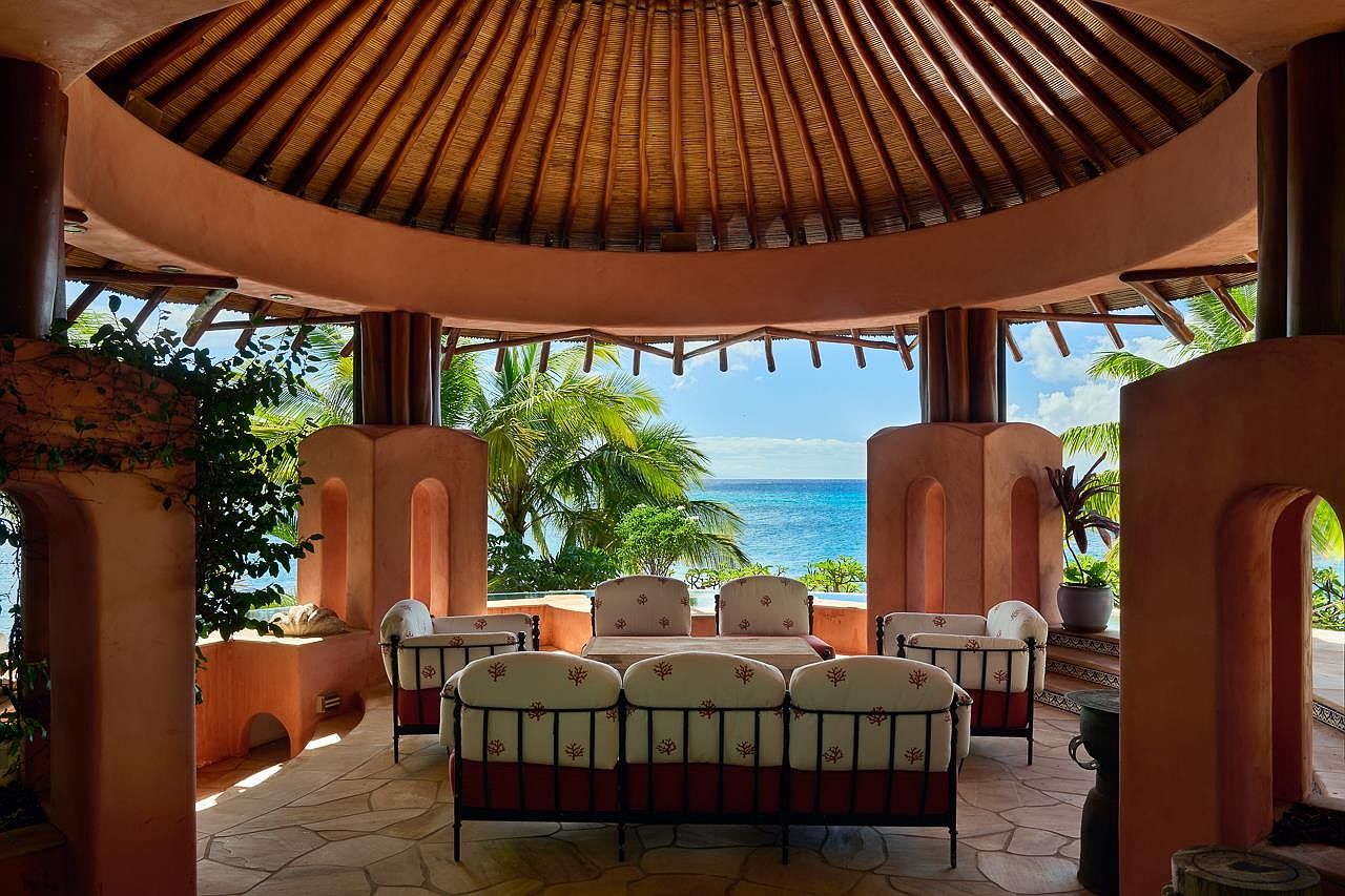 This image showcases a luxurious outdoor patio or balcony area with a stunning ocean view. The space is furnished with comfortable seating arrangements, featuring cushioned chairs and a sofa, all under a unique thatched roof structure supported by sturdy pillars. The design incorporates natural materials and colors, creating a serene and inviting atmosphere perfect for relaxation and enjoying the coastal scenery.