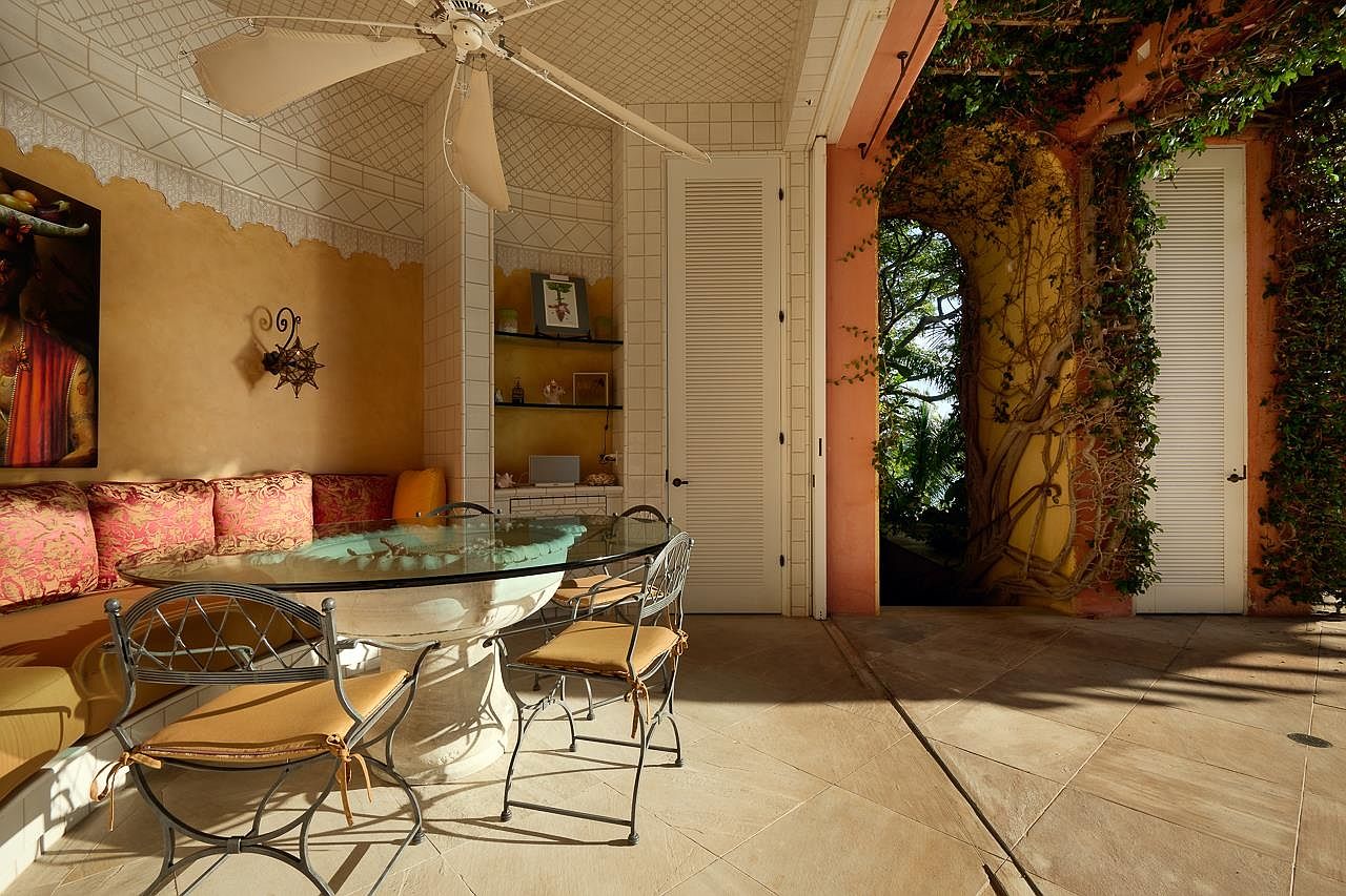 This image showcases a covered patio or balcony area, featuring a round glass-top dining table with wrought iron chairs and a built-in cushioned bench. The space is adorned with unique architectural details, including tiled walls and ceiling, and an arched entryway covered in vines, creating a charming and inviting outdoor dining experience. The flooring is tiled and the space is well lit.