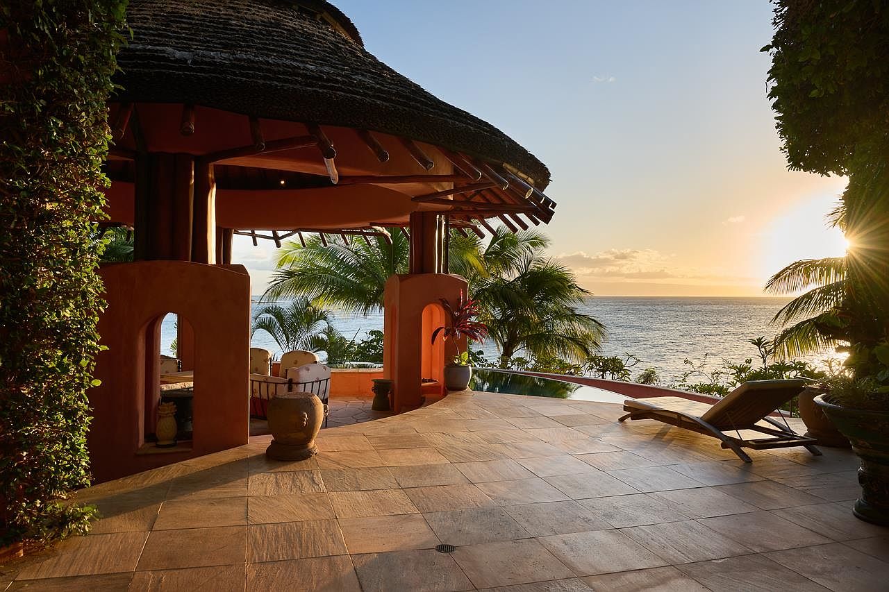 This image showcases a luxurious outdoor patio or deck area with a thatched-roof structure, overlooking a stunning ocean view at sunset. The space features tiled flooring, comfortable lounge seating, and lush tropical foliage, creating a serene and inviting atmosphere. The infinity pool blends seamlessly with the ocean horizon, enhancing the sense of tranquility and exclusivity.