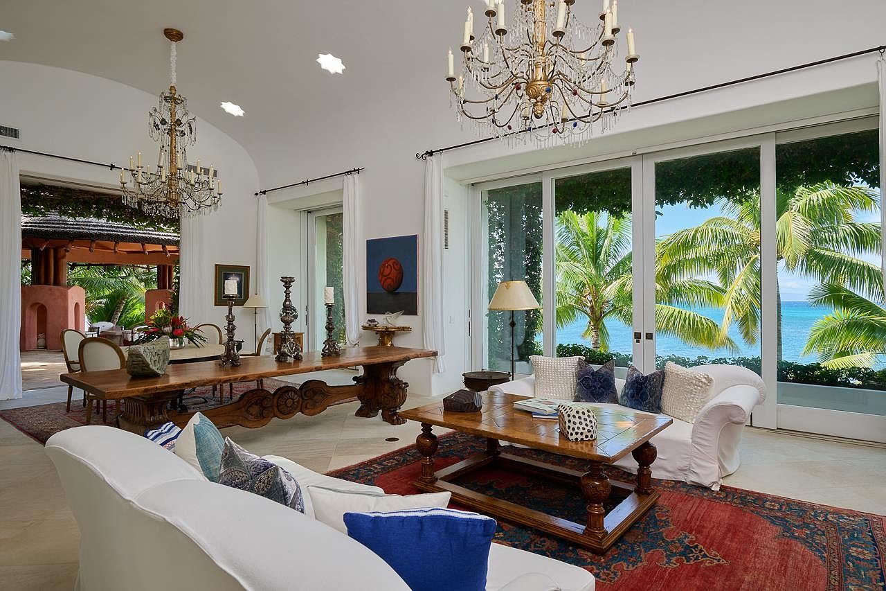 This is an interior shot of a luxurious living room that seamlessly blends indoor and outdoor living. The room features elegant chandeliers, white sofas with decorative pillows, and a large wooden coffee table atop an ornate rug. Sliding glass doors offer stunning views of palm trees and the ocean, creating a bright and airy atmosphere.