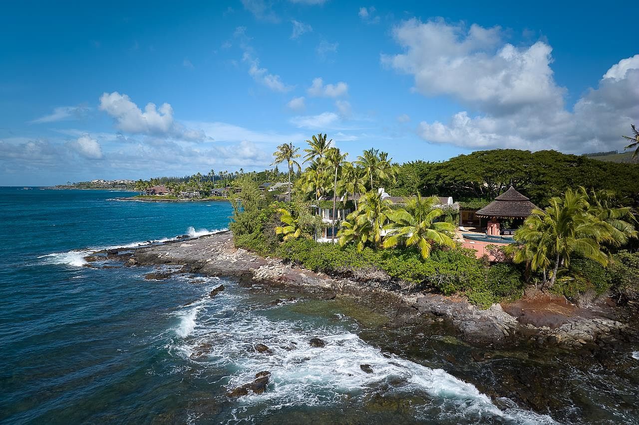 This aerial shot showcases a luxurious coastal property with a main house, a separate gazebo structure, and a swimming pool, all nestled amidst lush tropical vegetation. The property sits on a rocky coastline with the ocean waves crashing against the shore, creating a serene and exclusive atmosphere. The view extends to the coastline in the distance under a partly cloudy sky.