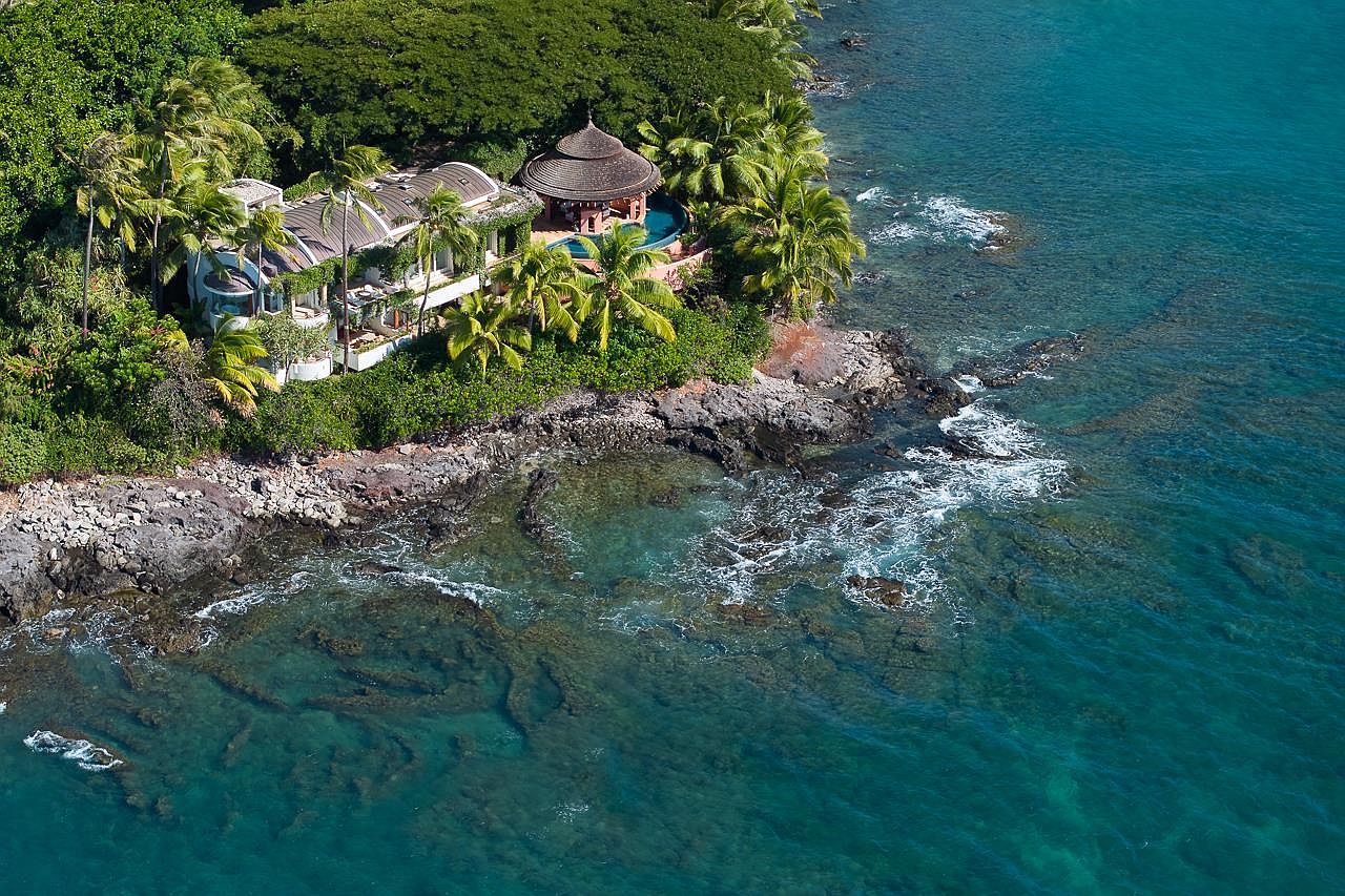 This aerial shot showcases a luxurious coastal property nestled amidst lush greenery and rocky shoreline. The residence features a unique architectural design with a rounded structure and a thatched-roof gazebo adjacent to a pool, blending seamlessly with the natural landscape. The turquoise waters surrounding the property create a serene and exclusive atmosphere, highlighting the property's prime waterfront location.