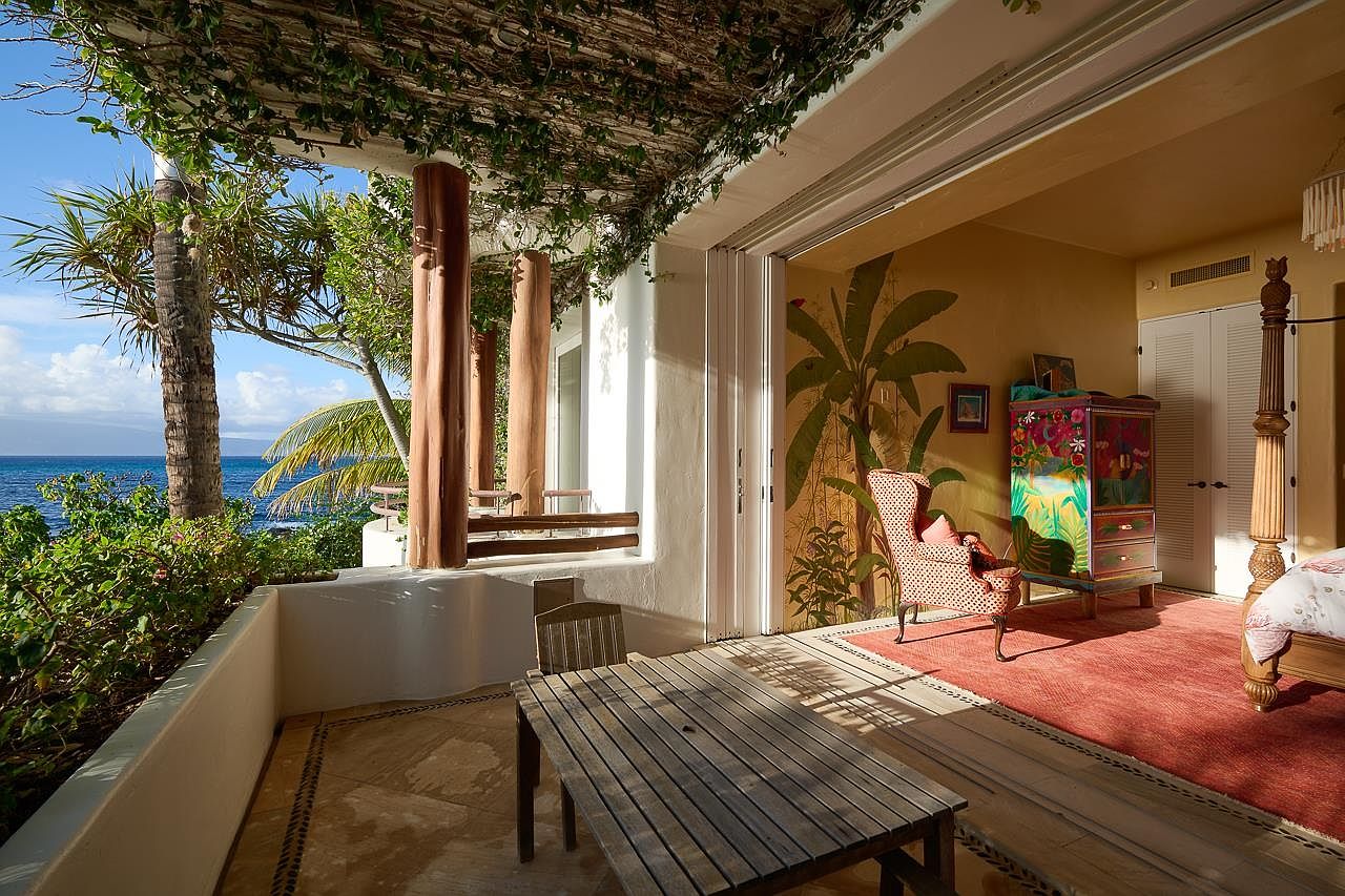 This image showcases a charming patio or balcony area with a wooden table and chairs, offering a serene view of the ocean. The space is partially covered by a vine-draped pergola, providing shade and a touch of nature. The interior of the adjacent room is visible, featuring a colorful armoire and a bed, suggesting a seamless indoor-outdoor living experience.
