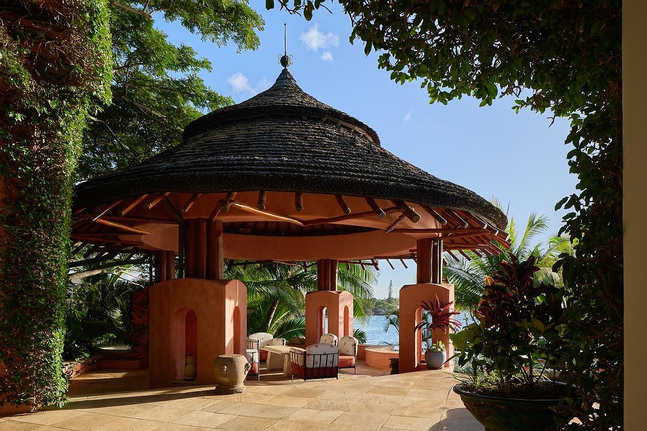 This image showcases a luxurious outdoor patio area featuring a thatched-roof gazebo with open archways and comfortable seating. The patio is paved with stone tiles and surrounded by lush tropical foliage, offering a serene view of the ocean in the background. The scene evokes a sense of relaxation and upscale outdoor living.