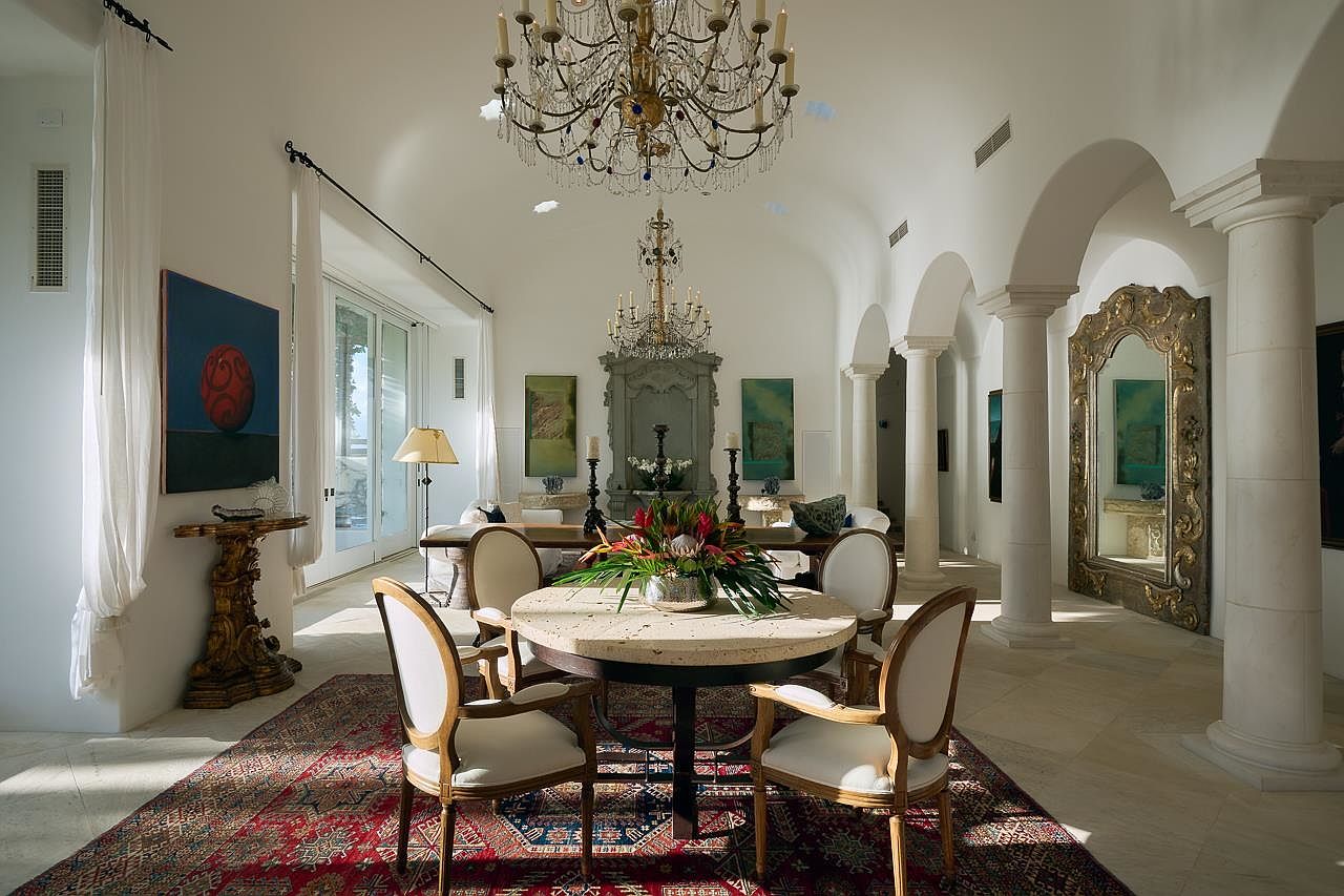 This is an elegant dining room featuring a round dining table with seating for four, set upon a large, ornate rug. The room is brightly lit, with natural light streaming in and supplemented by a grand chandelier. Architectural details include arched doorways and decorative columns, contributing to a luxurious and spacious atmosphere.