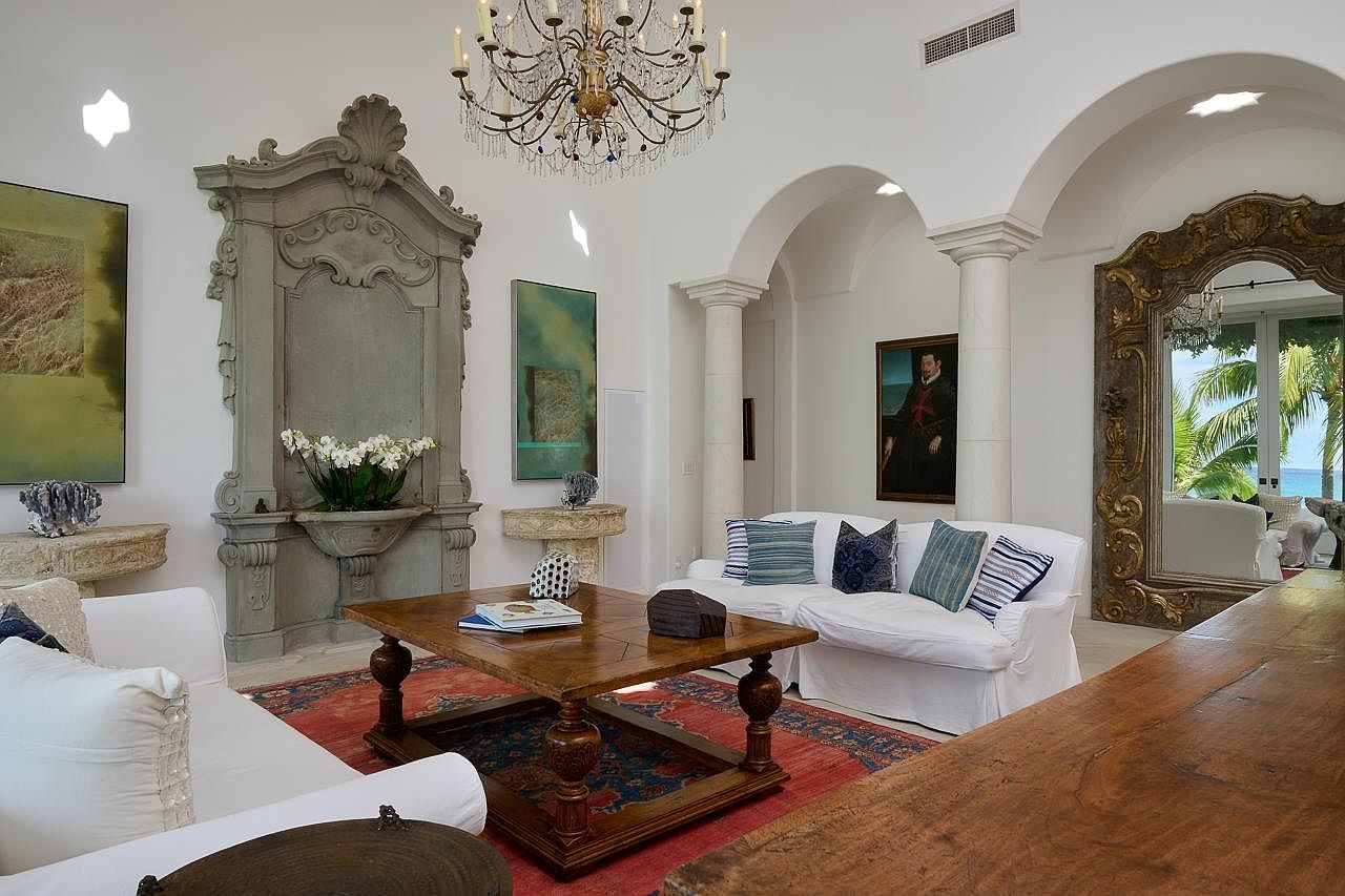 This is an interior shot of a luxurious living room featuring white sofas with decorative pillows, a large wooden coffee table on a red patterned rug, and an ornate stone fireplace. The room is bright with natural light and features arched doorways and a decorative chandelier, creating an elegant and inviting atmosphere. A large gilded mirror reflects the outdoor scenery, enhancing the sense of space and luxury.