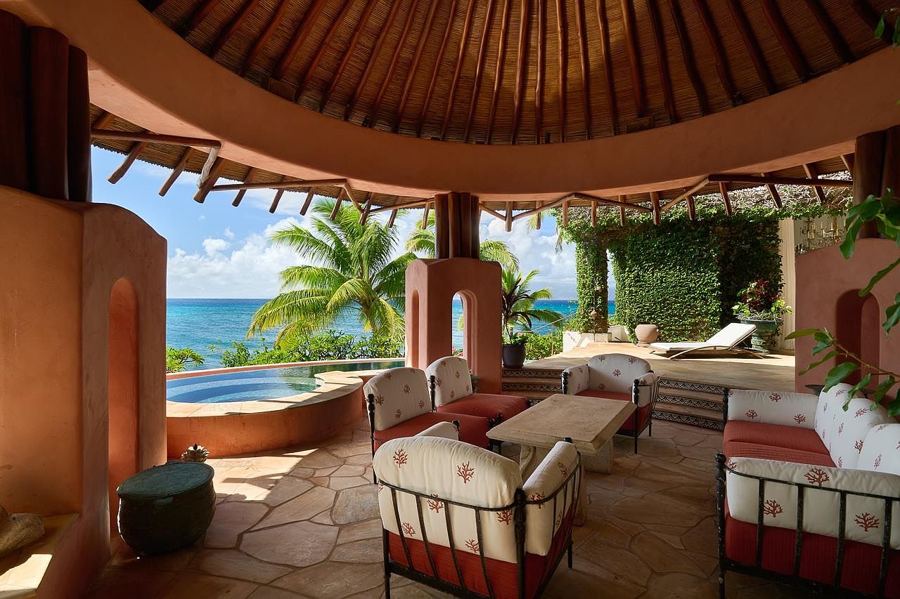 This image showcases a luxurious outdoor living space with a covered patio overlooking the ocean. The patio features comfortable seating arrangements, a stone table, and a small pool, all set against a backdrop of lush greenery and clear blue skies. The architectural design incorporates natural materials and a warm color palette, creating an inviting and relaxing atmosphere.