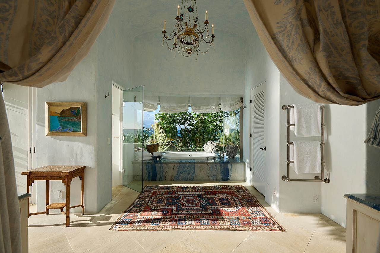 This is a luxurious primary bathroom featuring a freestanding tub with a view of lush greenery and the ocean. The room is decorated with a chandelier, a patterned rug, and elegant curtains that frame the space. The design creates a spa-like atmosphere with natural light and a serene ambiance.