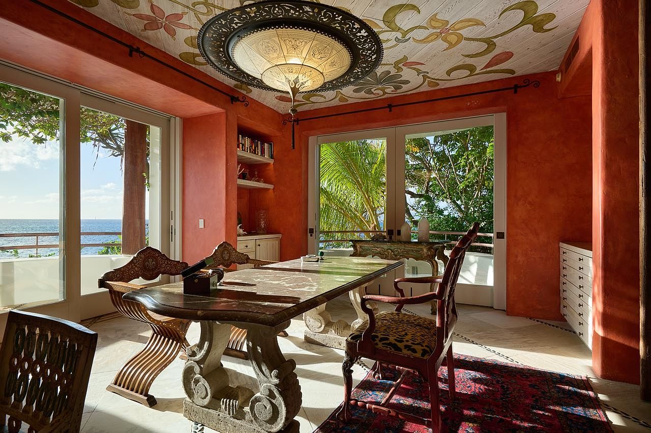 This dining room features a unique and ornate design with an orange color scheme. A large, decorative light fixture hangs from a ceiling adorned with floral patterns. The room includes a substantial dining table with carved stone legs, complemented by elegant chairs and a view of the ocean through large windows, creating a luxurious and inviting atmosphere.